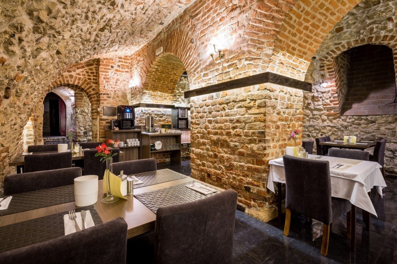 Przytulna restauracja w piwnicy z ceglanymi łukami, stołami nakrytymi i kwiatami w wazonach.