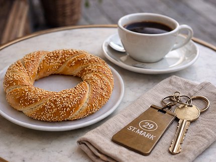 Sesame seed-covered circular bread on a white plate next to a cup of black coffee and a set of keys with a St. Mark room tag on a beige cloth napkin.