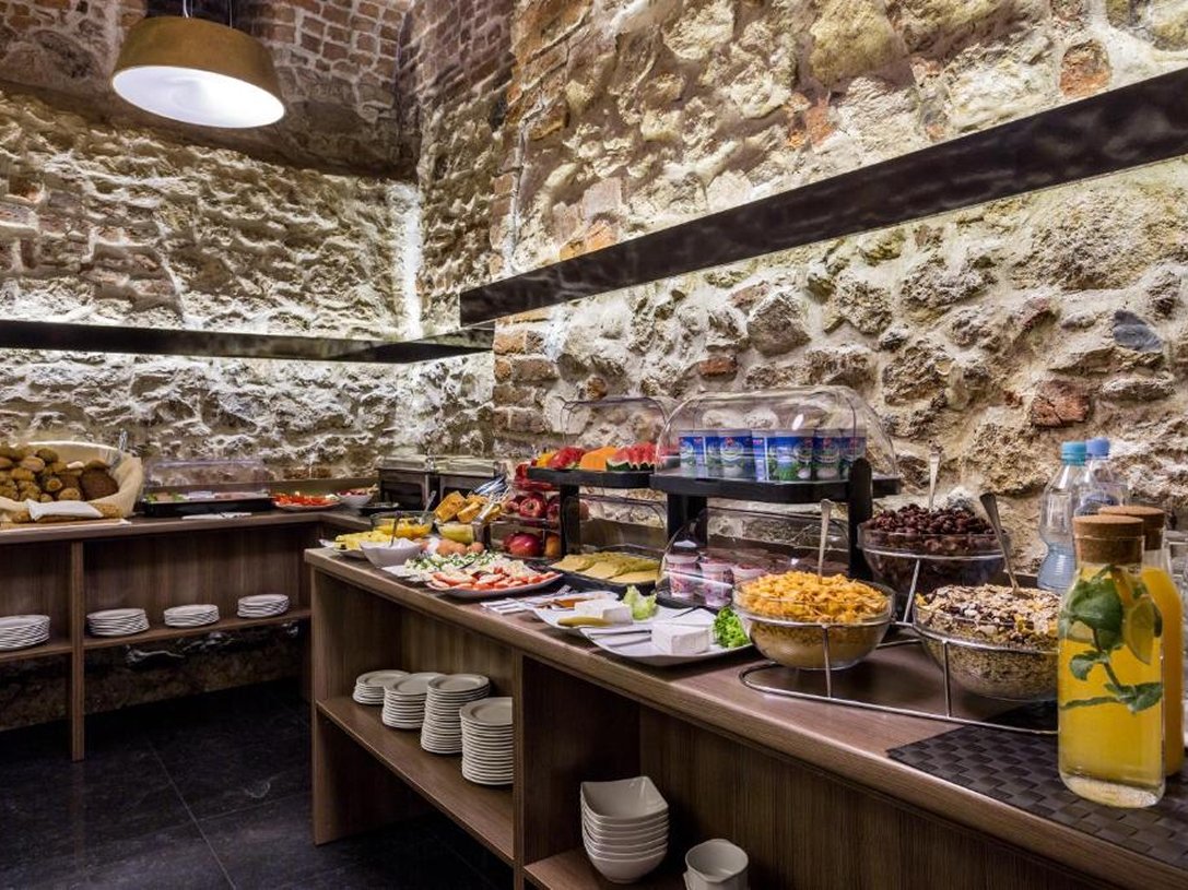 Bufet śniadaniowy z różnymi potrawami, takimi jak owoce, płatki śniadaniowe, sery, pieczywo i napoje w rustykalnym wnętrzu z kamiennymi ścianami.