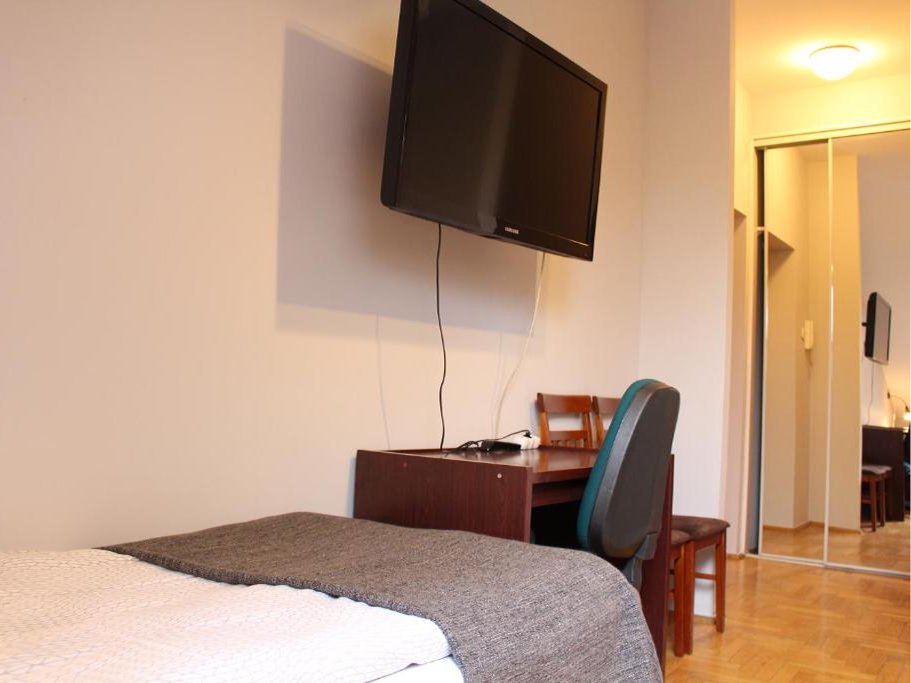 Pokój z łóżkiem przykrytym białą pościelą i szarą narzutą, biurkiem z dwoma krzesłami oraz telewizorem zamontowanym na ścianie.