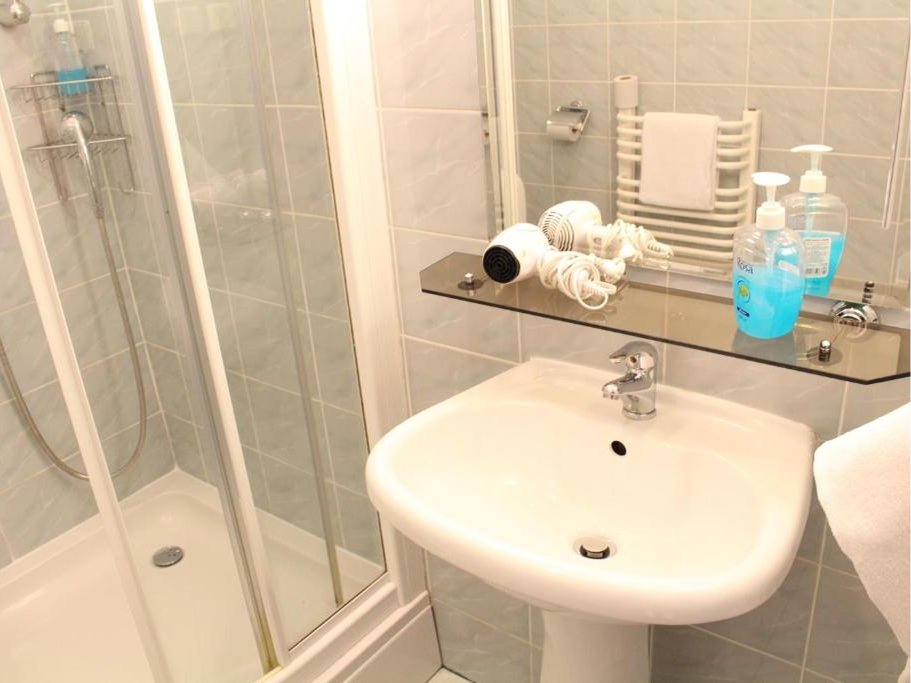 Łazienka z białą umywalką, półką z suszarką do włosów i mydłem w płynie, prysznicem oraz ręcznikiem na wieszaku.